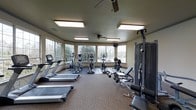 This fitness center offers a comprehensive workout environment designed to support a variety of exercise routines. The space is outfitted with multiple treadmills lining one side of the room, providing ample cardio options for users. A row of elliptical trainers and upright bikes occupies the central area, rounding out the cardio equipment selection. On the opposing side, a multi-station cable weight machine and a dedicated bench press setup cater to strength training needs. The room is finished with durable, low-pile carpet throughout, creating a practical and comfortable surface underfoot. Large, multi-pane windows wrap around the space, flooding the interior with natural light and offering pleasant views of the surrounding outdoor greenery. Two ceiling fans are mounted overhead to maintain airflow and comfort during workouts. Recessed flush lighting panels supplement the natural light, ensuring the space remains bright and well-lit at all hours. The neutral wall tones and functional layout contribute to a clean, focused atmosphere conducive to fitness. Overall, this single-floor facility balances cardio and strength equipment effectively within an airy, light-filled setting.
