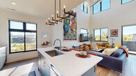This contemporary two-story home in Altoona, Wisconsin offers a thoughtfully designed layout spread across two well-proportioned floors. The main level opens into a combined kitchen and living area distinguished by soaring double-height ceilings and an abundance of natural light streaming through large, black-framed windows. The kitchen is outfitted with sleek grey cabinetry, stainless steel appliances, a mirrored tile backsplash, and a generous island with a white quartz countertop, undermount sink, and bar seating — anchored overhead by a sculptural candelabra-style pendant light. The open living room flows seamlessly from the kitchen, featuring light hardwood flooring and direct views to the outdoors. A dedicated laundry room houses a full-size side-by-side washer and dryer with overhead shelving. A modern bathroom on the main level features a floating wood vanity, quartz countertop, and a backlit oval mirror. The staircase with sleek cable railings leads to the upper level, where a spacious second living area enjoys views through large windows. The upper floor also includes a well-appointed bathroom, a generously sized walk-in closet with built-in shelving, and additional living space bathed in natural light.