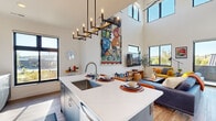 This contemporary two-story home in Altoona, Wisconsin offers a thoughtfully designed layout spread across two well-proportioned floors. The main level opens into a combined kitchen and living area distinguished by soaring double-height ceilings and an abundance of natural light streaming through large, black-framed windows. The kitchen is outfitted with sleek grey cabinetry, stainless steel appliances, a mirrored tile backsplash, and a generous island with a white quartz countertop, undermount sink, and bar seating — anchored overhead by a sculptural candelabra-style pendant light. The open living room flows seamlessly from the kitchen, featuring light hardwood flooring and direct views to the outdoors. A dedicated laundry room houses a full-size side-by-side washer and dryer with overhead shelving. A modern bathroom on the main level features a floating wood vanity, quartz countertop, and a backlit oval mirror. The staircase with sleek cable railings leads to the upper level, where a spacious second living area enjoys views through large windows. The upper floor also includes a well-appointed bathroom, a generously sized walk-in closet with built-in shelving, and additional living space bathed in natural light.