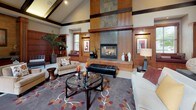 This refined single-floor space makes a striking first impression with a welcoming entrance foyer finished in warm-toned tile flooring accented by inlaid mosaic detailing. Craftsman-style wood columns with decorative stone inlays frame the entry corridor, leading visitors into the expansive main lounge area. The lounge is the architectural centerpiece of the space, boasting soaring vaulted ceilings with exposed dark wood beams and a dramatic skylight that fills the room with natural light. Floor-to-ceiling warm wood paneling wraps the walls, culminating in a grand stone and tile fireplace that serves as a commanding focal point. Built-in shelving with display niches flanks the fireplace, while a generous window seat offers a comfortable retreat with garden views. The lounge is thoughtfully furnished with plush seating arrangements, a decorative area rug, and a glass-top coffee table. A dedicated dining area provides an intimate setting centered around a round glass-top table with refined seating for four, accompanied by a sideboard and natural light filtering through draped windows. Throughout, the space exudes a warm, upscale ambiance defined by cohesive woodwork, quality finishes, and careful attention to architectural detail.