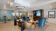 This thoughtfully designed single-floor space serves as a sophisticated communal hub, encompassing a grand entrance, a connecting hallway, and an expansive lounge area. The entrance greets visitors with large-format light tile flooring, floor-to-ceiling glass doors framed in matte black, and abundant natural light streaming in from exterior-facing windows. A central pedestal table topped with a stone surface and adorned with a decorative plant anchors the entry, while patterned area rugs add warmth underfoot.

The hallway transitions seamlessly into the main living area, lined with glass-paneled rooms featuring black-framed doors — offering visibility into a well-appointed office or meeting space and additional private rooms. A simple bench and framed artwork along the corridor contribute to an inviting, curated atmosphere.

The living lounge is the centerpiece of the space, designed for comfort and community. It features a dramatic dark marble-clad fireplace wall with a wall-mounted flat-screen television, complemented by an eclectic mix of blue velvet lounge chairs, gold accent seating, and warm wood coffee tables. A green accent wall with pendant lighting and landscape artwork defines a casual dining nook with high bistro tables and chairs. Ceiling-mounted HVAC units and recessed lighting ensure year-round comfort throughout this polished, modern space.