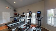 The fitness center at Southgate Landing is a thoughtfully designed, single-floor facility that provides residents with a comprehensive workout experience. The space is organized into distinct functional zones, allowing for a smooth flow between cardio training, strength work, and functional fitness. The cardio area features multiple treadmills, elliptical trainers, and a recumbent bike, each equipped with individual display screens for entertainment during workouts. Large windows with white blinds line the walls, filling the space with natural light and offering views of the surrounding greenery. The strength and free weight section includes a full rack of dumbbells, a weight bench, and a Life Fitness multi-station cable machine, complemented by large wall mirrors that enhance the sense of openness. Stability balls and additional functional training equipment round out the offerings. The flooring features a distinctive orange and tan patterned rubber surface that adds visual character while providing cushioning and durability. A built-in counter with cabinetry and a wall-mounted water fountain offer added convenience. The vaulted ceiling in portions of the space contributes to an airy, open ambiance, making this fitness center a well-appointed amenity for residents of Southgate Landing.