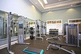 This fitness center is thoughtfully designed to support a comprehensive workout experience within a single, open-plan floor. The space immediately impresses with its soaring double-height ceilings, which create an airy, expansive atmosphere that enhances the sense of openness throughout. Large windows along the perimeter walls allow abundant natural light to pour in, offering pleasant views of the surrounding tree canopy and making the environment feel bright and inviting.

The cardio zone is well-appointed with a row of treadmills and stationary bikes, including Precor-branded equipment, providing a solid selection for cardiovascular training. Opposite the cardio area, a dedicated strength training section features a multi-functional cable and weight machine setup, catering to a variety of resistance exercises. Full-length mirrors line one wall, serving both a functional and aesthetic purpose — visually expanding the space while allowing users to monitor their form during workouts.

The flooring throughout is a durable, dark rubberized surface well-suited for a high-activity environment. Recessed and flush-panel ceiling lighting supplements the natural light, ensuring the space is well-lit at all hours. The overall ambiance is clean, modern, and functional, offering an accessible and motivating setting for fitness enthusiasts of all levels.