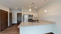 This thoughtfully designed single-floor residence offers a bright, open-concept layout that seamlessly connects the kitchen, dining, and living areas. The kitchen is a standout feature, showcasing a large granite-topped island with an undermount sink, crisp white upper cabinetry paired with dark lower cabinetry, and a full suite of stainless steel appliances including a refrigerator, range, and over-the-range microwave. Decorative pendant lights hang above the island, adding warmth to the contemporary aesthetic. Wood-look plank flooring flows throughout the main living areas, grounding the clean, neutral color palette. The living area is flooded with natural light through expansive floor-to-ceiling windows and a sliding glass door that opens to an outdoor view of lush tree canopy and a residential streetscape. The bedroom features plush carpeting, a ceiling fan, and a tall narrow window that brings in additional natural light. The bathroom is finished with neutral tile flooring, a walk-in shower, and a dark wood vanity topped with a white countertop and a large mirror. An in-unit stacked washer and dryer is also included. The overall ambiance is clean, contemporary, and move-in ready.