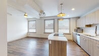 This single-floor unit blends modern design with industrial touches to create a bright, welcoming living space. The open-concept layout connects the kitchen and living area seamlessly, anchored by warm hardwood-style flooring that flows throughout. Exposed ductwork and structural elements add an industrial character to the otherwise clean, contemporary aesthetic.

The kitchen is a standout feature, showcasing light wood cabinetry that extends to the ceiling for ample storage, complemented by white quartz countertops and a textured tile backsplash. A central island with an integrated induction cooktop serves as both a prep area and a social focal point, while stainless steel appliances — including a full-size refrigerator, microwave, and dishwasher — complete the well-equipped space. Pendant lights with gold-toned shades hang above the island, adding warmth and style.

Multiple windows along the exterior walls fill the living area with natural light and offer views of the surrounding urban streetscape. The bedroom is generously proportioned, featuring its own windows and a ceiling fan for comfort.

The bathroom is finished with classic white subway tile in the tub and shower surround, a light wood vanity with white countertop, and hardwood-style flooring, maintaining the cohesive modern palette found throughout the unit.