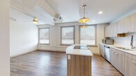 This single-floor unit blends modern design with industrial touches to create a bright, welcoming living space. The open-concept layout connects the kitchen and living area seamlessly, anchored by warm hardwood-style flooring that flows throughout. Exposed ductwork and structural elements add an industrial character to the otherwise clean, contemporary aesthetic.

The kitchen is a standout feature, showcasing light wood cabinetry that extends to the ceiling for ample storage, complemented by white quartz countertops and a textured tile backsplash. A central island with an integrated induction cooktop serves as both a prep area and a social focal point, while stainless steel appliances — including a full-size refrigerator, microwave, and dishwasher — complete the well-equipped space. Pendant lights with gold-toned shades hang above the island, adding warmth and style.

Multiple windows along the exterior walls fill the living area with natural light and offer views of the surrounding urban streetscape. The bedroom is generously proportioned, featuring its own windows and a ceiling fan for comfort.

The bathroom is finished with classic white subway tile in the tub and shower surround, a light wood vanity with white countertop, and hardwood-style flooring, maintaining the cohesive modern palette found throughout the unit.