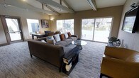 This single-story space showcases a striking architectural design highlighted by vaulted ceilings with exposed beam detailing and track lighting that adds both function and visual interest overhead. Large sliding glass doors and expansive windows line the walls, flooding the interior with generous natural light while offering clear views of the outdoor surroundings, including mature trees and an open exterior area.

The main living area is anchored by a substantial L-shaped sofa in deep charcoal, accented with a mix of neutral and rust-toned throw pillows that add warmth and texture. A sleek, dark coffee table and a glass-topped side table complement the seating arrangement, while a patterned area rug grounds the space and adds dimension underfoot.

In the background, a dining or game area is visible, featuring a dark wood table surrounded by upholstered chairs, suggesting the space is well-suited for both lounging and social gatherings. Wall art and a warm sconce light contribute to the inviting ambiance. The overall aesthetic blends contemporary furnishings with architectural character, resulting in a comfortable and visually cohesive environment.