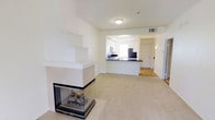 This single-floor residence offers a clean and open layout bathed in a neutral, light-filled palette throughout. The living area serves as the heart of the space, anchored by a striking three-sided corner fireplace with a sleek black metal frame and stone surround, adding both warmth and a modern focal point to the room. Plush carpeting covers the living area, contributing to a comfortable and inviting atmosphere.

The kitchen is accessible directly from the living space through an arched pass-through opening, creating a connected yet distinct area. It features white cabinetry, dark countertops, and a breakfast bar overhang that provides casual seating. A window above the sink allows natural light to brighten the workspace. The kitchen's design is both functional and aesthetically cohesive with the rest of the home.

Adjacent to the kitchen, the entryway is finished with light hardwood-style flooring and includes a full-height entry door with sidelight, offering a welcoming transition into the home. Visible HVAC vents and flush-mount lighting are integrated cleanly into the white ceilings. The consistent white walls and trim throughout create a bright, airy feel, making the space feel open and well-proportioned across all areas of the floor plan.