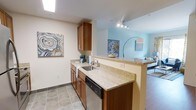 This single-story residence features a thoughtfully designed open-concept layout that maximizes its footprint. The kitchen is a standout feature, showcasing warm medium-toned wood cabinetry paired with speckled granite countertops that extend along both the perimeter and a peninsula breakfast bar. The peninsula creates a natural division between the kitchen and living area while maintaining an open sightline throughout the space. A stainless steel sink, dishwasher, and a skylight overhead lend both functionality and brightness to the kitchen. Tile flooring grounds the kitchen area, providing a durable and clean aesthetic. The adjoining living space is painted in a soft blue-green hue, creating a calm and inviting atmosphere. Track lighting in the living area provides adjustable illumination, complementing the overall modern sensibility of the unit. Neutral wall tones in the kitchen contrast gently with the accent color of the living space, adding visual depth. Decorative wall art adds personality throughout. The layout flows cohesively from room to room, making the space feel connected and efficient.