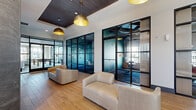 This thoughtfully designed single-floor space serves as a multifunctional lobby and communal lounge area, offering a generous footprint that balances open gathering areas with enclosed private spaces. The layout centers around an open lounge furnished with contemporary cream upholstered seating, set atop wood-look tile flooring that runs throughout the space, lending warmth and continuity.

Flanking the lounge are floor-to-ceiling black steel-framed glass partition walls enclosing private conference or meeting rooms, accented with teal-tinted glass panels that add a striking pop of color. These transparent enclosures maintain visual connectivity while providing acoustic separation. Overhead, a combination of recessed lighting and statement gold dome pendant fixtures illuminates the open areas, while a richly stained dark wood ceiling panel adds architectural contrast.

A dramatic accent wall clad in deep slate-toned marbled panels anchors one end of the space, housing a stainless steel elevator and a flush-panel door. Nearby, a built-in leather banquette with individual white-topped work surfaces sits beneath an ornate geometric metal screen partition, creating a stylish café-style nook. A wall-mounted display screen and a shiplap wood accent wall complete the space, adding functional and aesthetic variety throughout.
