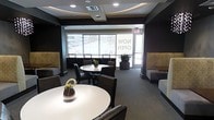 This single-floor space presents a polished, contemporary lounge environment designed for comfort and social interaction. The interior is defined by a sophisticated dark color palette, with deep charcoal walls and dark furniture creating a sleek, modern aesthetic. The ceiling features a distinctive recessed oval tray detail with integrated lighting, adding architectural interest and a refined focal point overhead.

The main area is furnished with multiple round white tables surrounded by dark upholstered chairs, encouraging a casual yet professional atmosphere. Built-in booth-style seating lines the perimeter walls, upholstered in neutral tones with textured tile or mosaic accent panels behind them, adding a layer of visual depth and warmth to the darker surroundings.

Large floor-to-ceiling windows span the far wall, flooding the space with natural light and providing views of the surrounding urban landscape. A glass-paneled door offers direct access to an exterior area. Lush potted plants are strategically placed near the windows, introducing natural elements that complement the modern design. Recessed ceiling lighting and coordinated décor accents complete the well-appointed, inviting atmosphere throughout.
