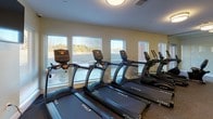 This well-equipped fitness center in Castle Shannon, PA offers a comprehensive workout environment designed to support a variety of exercise routines. The space is thoughtfully laid out across a single floor, with designated zones for both cardio and strength training. A row of True-brand treadmills lines one wall, positioned beneath a bank of large windows that flood the room with natural light and provide pleasant outdoor views. An elliptical machine rounds out the cardio section, while a wall-mounted flat-screen TV adds entertainment during workouts. The strength training area features a cable machine, an adjustable weight bench, a full dumbbell rack with a wide range of weights, a medicine ball tower, and stability balls, catering to users of all fitness levels. The flooring throughout is rubber gym matting, providing comfort and durability underfoot. Flush-mounted ceiling lights ensure bright, even illumination throughout the space. A glass partition wall opens the fitness center toward a corridor, enhancing the sense of openness and connectivity within the building. The overall ambiance is clean, modern, and functional, making it a well-rounded facility for daily fitness needs.