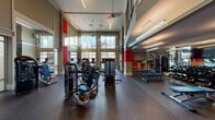 This fitness center is designed to deliver a full-featured workout experience within a striking architectural setting. The space is defined by soaring double-height ceilings and expansive floor-to-ceiling windows that flood the interior with natural light and offer open views of the surrounding community. Bold red accent panels punctuate the neutral taupe and gray walls, adding energy and visual contrast throughout.

The main workout floor is finished with durable rubber flooring and is organized into distinct training zones. A dedicated cardio area lines the windowed wall, featuring a row of Matrix treadmills and additional cardio machines positioned to take advantage of the outdoor views. The strength training zone is well-appointed with a wide selection of selectorized weight machines and cable systems, providing comprehensive options for both upper and lower body training. A free weights area includes multiple weight benches and a full rack of dumbbells, complemented by large wall mirrors and stylish pendant lighting that give this zone a more intimate, focused atmosphere.

A spiral staircase adds an architectural focal point, connecting to an upper-level mezzanine. The combination of abundant natural light, high ceilings, modern equipment, and thoughtful zoning creates a motivating and functional environment suited to a wide range of fitness routines.