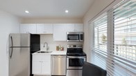 This single-floor space includes a well-appointed kitchen that serves as a functional and visually clean area. The kitchen is outfitted with a full suite of stainless steel appliances, including a refrigerator, electric range, over-the-range microwave, and dishwasher, offering a cohesive and modern aesthetic. White shaker-style cabinetry lines both the upper and lower walls, providing ample storage while contributing to a bright, airy feel throughout the space. The countertops are light-colored, complementing the overall neutral palette of white walls and warm-toned flooring. A stainless steel sink with a modern faucet is neatly integrated beneath the cabinetry. A large window fitted with horizontal blinds allows natural light to filter into the kitchen, enhancing the sense of openness. Recessed ceiling lighting provides additional illumination. The layout is efficiently designed, making practical use of the available space. The overall ambiance is clean, contemporary, and functional, with a neutral color scheme that offers a versatile foundation for personalization.