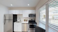 This single-floor space includes a well-appointed kitchen that serves as a functional and visually clean area. The kitchen is outfitted with a full suite of stainless steel appliances, including a refrigerator, electric range, over-the-range microwave, and dishwasher, offering a cohesive and modern aesthetic. White shaker-style cabinetry lines both the upper and lower walls, providing ample storage while contributing to a bright, airy feel throughout the space. The countertops are light-colored, complementing the overall neutral palette of white walls and warm-toned flooring. A stainless steel sink with a modern faucet is neatly integrated beneath the cabinetry. A large window fitted with horizontal blinds allows natural light to filter into the kitchen, enhancing the sense of openness. Recessed ceiling lighting provides additional illumination. The layout is efficiently designed, making practical use of the available space. The overall ambiance is clean, contemporary, and functional, with a neutral color scheme that offers a versatile foundation for personalization.