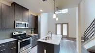 This contemporary two-story residence showcases a thoughtfully designed open-concept layout with high-end finishes throughout. The kitchen serves as a central highlight, featuring dark espresso-toned cabinetry extending to ceiling height, light granite countertops, a mosaic tile backsplash, and a full suite of stainless steel appliances including a refrigerator, range, and built-in microwave. A central kitchen island with an undermount sink and pendant lighting above provides both additional workspace and a natural gathering point. The living area benefits from dramatic double-height ceilings and clerestory windows that draw in abundant natural light, creating an airy, open ambiance. A sleek staircase with a modern metal railing leads to the upper level, which overlooks the main floor below through an open loft-style railing. The upper-level bedroom is generously sized with plush carpeting and a ceiling fan, while the walk-in closet offers wrap-around hanging rods and built-in shelving for ample storage. The bathroom is finished with a dark wood vanity, granite countertop, a large mirror, and a full soaking tub with shower. Light wood-look flooring runs throughout the main level, tying the modern aesthetic together cohesively.
