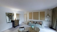This single-floor space presents a thoughtfully arranged living area with a distinct classic aesthetic. The room is anchored by a tufted sofa upholstered in a silvery-blue damask fabric, complemented by a round glass-top coffee table that adds a refined touch to the seating area. A cushioned accent chair provides additional seating, contributing to the room's comfortable and welcoming layout.

A prominent design feature is the set of four gold lattice wall panels mounted along one wall, creating a bold decorative focal point that lends warmth and visual texture to the otherwise neutral space. The walls are finished in a clean, light tone, and the smooth white ceiling enhances the sense of openness throughout the room.

To one side, a dark-finished dresser with gold hardware is positioned beneath a framed mirror, suggesting a multifunctional use of the space. A table lamp with a classic shade provides ambient lighting, adding to the room's warm, layered character. The carpeted flooring in a neutral hue ties the space together, creating a cohesive and inviting interior atmosphere throughout.