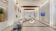 Located at 1350 S Dixie Hwy in Coral Gables, Florida, this single-floor lobby space exemplifies upscale contemporary design with a refined, welcoming atmosphere. The entrance is framed by floor-to-ceiling glass doors and windows, flooding the interior with natural light and offering views of lush greenery outside. Soaring ceilings with warm wood beam accents and recessed lighting create a dramatic sense of height and openness. The flooring features a distinctive geometric patterned tile that anchors the central lounge area, bordered by smooth light wood and stone surfaces. A sculptural reception desk sits against a striking accent wall adorned with flowing linear artwork, serving as a refined focal point. The lounge area is furnished with a generous curved sectional sofa and an organic-shaped white coffee table, encouraging a relaxed yet polished environment. Along one wall, a series of illuminated built-in niches display curated decorative objects and greenery, adding texture and visual depth. Floor-length white drapery panels line portions of the walls, softening the architecture and contributing to the space's serene, airy ambiance. A wall-mounted display screen provides informational content for visitors. The overall design balances modern minimalism with warm natural materials, creating an inviting and sophisticated arrival experience.