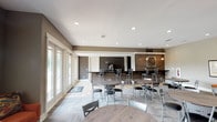 This single-floor gathering space offers a contemporary and well-appointed interior designed for communal use and social interaction. The open-concept layout accommodates multiple dining groupings, with round and rectangular tables paired with sleek metal chairs arranged throughout the room, creating a welcoming and functional environment.

The space is defined by a thoughtful blend of modern and rustic design elements. A standout feature is the wood-paneled accent wall at the rear, adorned with a large decorative clock and flanked by warm pendant lighting that adds an industrial charm. Adjacent to this wall is a bar-height counter area, ideal for casual seating or service, with a dark contrasting backdrop that anchors the space visually.

Flooring throughout combines light-toned tile and herringbone-patterned wood-look panels, adding texture and visual depth to the open floor plan. Recessed ceiling lighting provides consistent illumination across the space, complementing the natural light that pours in through a full wall of large windows and glass-paned doors along one side. These expansive windows offer views of outdoor greenery and allow abundant daylight to fill the interior, creating a bright and airy atmosphere. The neutral color palette of whites, grays, and warm wood tones ties the design together cohesively, resulting in a polished and inviting space.