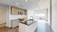 Located in Houston's sought-after Midtown area, this single-floor residence at 3815 Eastside St blends industrial character with modern design. The open-concept living and dining area is anchored by striking exposed concrete ceilings and structural columns, lending a loft-like aesthetic. Large floor-to-ceiling windows flood the space with abundant natural light while framing expansive views of the surrounding Houston skyline and cityscape, creating a bright and airy ambiance.

The well-appointed kitchen features light wood cabinetry, dark granite countertops, a subway tile backsplash, and a full suite of stainless steel appliances including a side-by-side refrigerator, electric range, built-in microwave, and dishwasher. A central island with an undermount sink adds both prep space and a natural division between the kitchen and living areas. Warm wood-tone flooring flows throughout the main living space, transitioning to neutral tile in the hallway corridor.

The hallway connects to a bathroom and a dedicated closet, offering functional storage solutions. Clean white walls throughout keep the space feeling open and versatile. The thoughtful layout and high-end finishes make this a well-designed urban residence ideal for modern living in Houston.