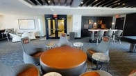 This contemporary lounge space showcases a striking interior design defined by bold contrasts and modern sophistication. The ceiling is a standout architectural feature, with dark exposed beams set against a white background, punctuated by recessed and track lighting that creates a dynamic, layered illumination throughout the room.

The space is organized into distinct seating zones, anchored by a large, circular orange leather ottoman that serves as a central gathering point. Surrounding it are curved, high-backed upholstered chairs in neutral grey tones, complemented by small accent tables featuring polished stone or agate tops. This central arrangement encourages social interaction while maintaining a stylish aesthetic.

Along one wall, a wall-mounted flat-screen television is flanked by built-in shelving displaying decorative vessels, with dark wall panels providing a dramatic backdrop. A linear fireplace is also visible, adding a warm, inviting element to the otherwise cool, modern palette.

Toward the back of the space, a bar-height table with tall chairs offers an alternative seating option, alongside framed artwork enhancing the refined atmosphere. The patterned carpet grounds the room, while glass-paneled doors connect the lounge to adjacent areas, contributing to an open, interconnected flow.