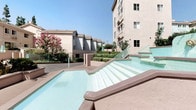 This property is part of a thoughtfully designed multi-building complex that blends functional architecture with inviting outdoor spaces. The exterior presents a clean, stucco-finished facade in warm earth tones, complemented by accent staircases finished in a distinctive light teal, adding a touch of color to the overall aesthetic.

The outdoor common areas are a notable highlight of the complex. A prominent water feature with shallow, tiered steps creates a visually engaging focal point, reflecting natural light and contributing to a relaxed, resort-like atmosphere. Surrounding greenery, including neatly trimmed hedges and mature flowering shrubs, enhances the sense of a well-cared-for environment.

The streetside view reveals a tree-lined boulevard with tall, mature trees providing generous shade and a lush canopy that adds privacy and curb appeal. The wide paved walkway at the entrance uses warm-toned brick pavers, creating a welcoming approach to the property.

The complex benefits from an abundance of natural light throughout its outdoor spaces, with clear blue skies and an open layout that promotes a bright and airy ambiance. The combination of architectural details and landscaped grounds makes this a visually appealing and comfortable setting.