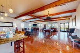 This single-story space in Visalia, California, showcases a rich Spanish Colonial aesthetic that carries consistently throughout its generous, open floor plan. Prominent exposed wood ceiling beams draw the eye upward, complementing the warm, polished terracotta tile flooring that runs seamlessly across the entire space. Wrought iron chandeliers and pendant lights reinforce the rustic yet refined character of the interior.

The expansive main hall serves multiple functions, offering distinct zones for dining, lounging, and recreation. A large dining table surrounded by ornate, high-backed leather chairs anchors one section, while a full-sized billiard table with carved wood legs serves as a centerpiece for the game area. A seating nook near a decorative tile-surround fireplace provides a more intimate setting, accompanied by leather armchairs, a wall-mounted television, and a built-in cue rack.

The kitchen features white cabinetry, granite countertops, stainless steel appliances including a double oven range and refrigerator, and a large island with bar seating. Multiple windows throughout the space invite natural light and offer views of the surrounding landscape, enhancing the warm, inviting ambiance of this versatile gathering space.