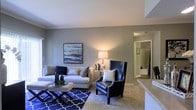 This single-floor residence offers a thoughtfully designed living area that serves as the centerpiece of the space. The living room showcases a neutral gray color palette on the walls, complemented by plush carpeted flooring that adds warmth underfoot. Crown molding accents the upper walls, lending a refined architectural touch to the room.

The space is furnished with a generous light gray sectional sofa paired with a bold navy leather wingback accent chair, creating a striking contrast that anchors the room. A patterned navy and white area rug adds visual interest beneath a sleek geometric coffee table. Ambient lighting is provided by a classic table lamp, casting a warm glow throughout the room.

A framed abstract artwork in blues and golds adorns the wall, serving as a focal point above the sofa. The layout flows naturally toward a hallway visible in the background, which appears to lead to additional rooms, including what looks to be a bathroom with tiled finishes. A countertop edge visible at the frame's right side suggests proximity to a kitchen or bar area, hinting at an open and connected floor plan throughout the residence.