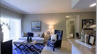 This single-floor residence offers a thoughtfully designed living area that serves as the centerpiece of the space. The living room showcases a neutral gray color palette on the walls, complemented by plush carpeted flooring that adds warmth underfoot. Crown molding accents the upper walls, lending a refined architectural touch to the room.

The space is furnished with a generous light gray sectional sofa paired with a bold navy leather wingback accent chair, creating a striking contrast that anchors the room. A patterned navy and white area rug adds visual interest beneath a sleek geometric coffee table. Ambient lighting is provided by a classic table lamp, casting a warm glow throughout the room.

A framed abstract artwork in blues and golds adorns the wall, serving as a focal point above the sofa. The layout flows naturally toward a hallway visible in the background, which appears to lead to additional rooms, including what looks to be a bathroom with tiled finishes. A countertop edge visible at the frame's right side suggests proximity to a kitchen or bar area, hinting at an open and connected floor plan throughout the residence.