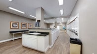This shared laundry facility is thoughtfully designed to provide a clean, functional, and comfortable experience for its users. The single-floor layout is open and spacious, with light wood-tone flooring and neutral gray walls that create a bright, welcoming atmosphere. Overhead, recessed fluorescent panel lighting ensures the entire space is well-illuminated throughout the day.

At the heart of the facility is a centrally positioned island unit featuring white cabinetry and a dark countertop with an integrated utility sink, offering a convenient handwashing and pre-treatment station. A paper towel dispenser is mounted directly on the island's column for easy access. Flanking the island is an extensive row of top-load washing machines, providing ample capacity for multiple users simultaneously. Stacked dryer units are also visible along one wall, maximizing the facility's laundry throughput.

Along one side of the room, a wall of individual storage lockers offers secure personal storage for residents. A long folding and sorting bench runs the length of another wall, providing generous counter space for managing laundry. Decorative framed artwork with coastal and nature-themed prints adds a homey touch to the utilitarian space. A window at the far end of the room allows natural light to filter in, further enhancing the open feel of the facility.