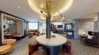 This single-floor amenity lounge is thoughtfully designed to offer a variety of functional and social environments within one cohesive space. At its centerpiece sits a striking circular table anchored by a substantial cylindrical column, topped by an eye-catching sculptural ceiling feature with warm gold tones and radiating brass light fixtures that draw the eye upward. Textured carpeting with an organic pattern grounds the space throughout.

Distinct seating zones cater to different needs: an open lounge area features rounded swivel chairs and soft seating arranged for casual conversation, while an intimate nook with a bold terracotta-orange accent wall showcases decorative floral wall art and plush, pillow-laden sofas around a round wood coffee table. An adjacent curved banquette area with a sculptural layered ceiling and pendant globe lighting offers a more enclosed, comfortable retreat complete with a game set.

Separated by sliding glass doors with gold hardware, a private conference or dining area provides a focused setting with a dark table and vibrant burnt-orange chairs alongside open shelving. Gray wood-look tile cladding defines a dedicated workspace alcove housing printing and copying equipment. The bar and lounge area visible beyond glass partitions features pendant lighting and additional seating, completing this well-appointed, design-forward space.