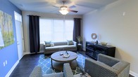 This single-floor space offers a thoughtfully designed interior with a focus on comfort and modern style. The living room serves as the centerpiece, showcasing dark hardwood-style flooring that contrasts elegantly with the light-toned walls. A large window dressed with dark curtains and white blinds allows an abundance of natural light to flow into the room, creating a bright and welcoming atmosphere. A ceiling fan with an integrated light fixture is centrally positioned overhead, providing both airflow and additional illumination. The living area is furnished with a neutral-toned sofa accented by patterned throw pillows, complemented by a round wooden coffee table resting on a teal geometric-patterned area rug. Additional seating in the form of patterned accent chairs adds depth and visual interest to the layout. A dark media console along one wall provides practical storage and display space, completing the room's balanced aesthetic. The overall ambiance is contemporary and cohesive, with a careful blend of textures, patterns, and tones that create an inviting and functional living environment.