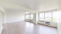 This spacious 2-bedroom, 2-bathroom residence is thoughtfully laid out across a single floor, offering a comfortable and functional living environment. The expansive living room is a standout feature, with multiple large windows spanning nearly the full width of the room, framing sweeping views of a river and lush treetop canopy. Light hardwood-style flooring, crisp white walls, and baseboard heating units run beneath the windows, contributing to a clean, airy aesthetic throughout the common areas.

The kitchen is galley-style and well-equipped, featuring dark cabinetry, dark granite countertops, black appliances including a range and built-in microwave, and a stainless steel refrigerator. Light wood flooring carries through this space, providing contrast to the darker finishes.

The generously sized primary bedroom features carpeting, a large window with outdoor views, a closet, and built-in shelving, offering ample storage. The second bedroom is similarly carpeted with good natural light. A separate dining room provides dedicated space for meals. Both bathrooms are finished with white tile and clean, minimal fixtures. A hallway connects the rooms efficiently, completing this well-organized residence.