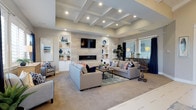 This elegant single-floor residence centers around a generously sized great room that immediately impresses with its architectural sophistication. The room's standout feature is a striking coffered ceiling with recessed lighting, adding depth and a refined character to the space. A sleek, linear gas fireplace serves as the focal point of the main wall, clad in light-toned tile that extends from floor to ceiling and is flanked by symmetrical built-in shelving units with cabinet storage below — ideal for both display and practical use. A wall-mounted flat-screen television is integrated seamlessly above the fireplace. The open layout accommodates generous seating arrangements with plush neutral sofas accented by vibrant navy and coral throw pillows, resting atop a layered area rug over soft carpet flooring. Large windows dressed in navy drapes allow ample natural light to fill the room while offering views to the exterior. The overall ambiance is warm and contemporary, with a cohesive palette of soft neutrals, whites, and navy blue accents that lend the space a coastal-inspired elegance. The thoughtful design balances comfort with style, making this great room an inviting setting for both relaxed living and entertaining.