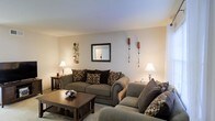 This two-floor space offers a comfortable and well-appointed living environment with a neutral, earthy color palette that creates a welcoming ambiance. The living area is a central feature, showcasing plush, dark-toned sofas adorned with patterned throw pillows in warm browns and taupes. A rectangular wooden coffee table anchors the seating arrangement, providing both functionality and a grounding visual element. The walls are painted in soft, light tones that enhance the brightness of the room, complemented by warm ambient lighting from elegantly styled table lamps positioned on either side of the space. Decorative wall sconces and a framed mirror add visual interest and depth to the room. The flooring transitions between carpet in the living area and what appears to be harder flooring near the entryway, adding textural variety throughout. The overall design aesthetic is cohesive and understated, balancing practicality with comfort. The layout across two floors suggests additional living, sleeping, or utility spaces beyond what is immediately visible, offering a well-rounded and functional home environment.