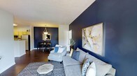 This single-floor space makes a strong design statement through its use of bold navy blue accent walls, which anchor both the living and dining areas with a sense of depth and sophistication. The open-concept layout allows the living room and dining area to flow seamlessly into one another, creating a cohesive and inviting atmosphere.

The living area is centered around a comfortable grey sectional sofa adorned with an assortment of blue, grey, and yellow throw pillows. A large abstract canvas painting in gold, white, and grey tones hangs prominently on the navy feature wall, serving as a striking focal point. A geometric white coffee table and a patterned area rug add texture and visual interest to the space.

Transitioning into the dining area, a warm-toned pendant light fixture hangs above a dark wood dining table surrounded by matching chairs. A mirror on the navy wall reflects light and enhances the sense of space. Dark hardwood flooring runs throughout, grounding the design and complementing the bold color palette. The overall ambiance is modern, curated, and welcoming.