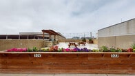 This rooftop-level space combines functional open-air parking with a thoughtfully designed outdoor patio area, offering an appealing urban retreat above the city. The parking area is clearly marked with yellow-painted stalls and provides open views of the surrounding downtown skyline, including notable historic and modern buildings that frame the horizon. Adjacent to the parking, the patio area is a standout feature, anchored by a warm wood pergola and strung with overhead bistro lights that span the space to create an inviting atmosphere. Raised wooden planter beds, including one prominently numbered 532, are filled with vibrant seasonal flowers and greenery, adding color and life to the rooftop setting. Picnic-style seating is arranged across the stone-tiled patio surface, making the area well-suited for outdoor gatherings. A stone-accented outdoor kitchen or grill station is tucked beneath the pergola, further enhancing the entertaining potential of the space. Additional cedar planter boxes throughout contribute to the garden-like character of the patio. The combination of open sky, city views, lush plantings, and comfortable amenities makes this rooftop an exceptionally functional and visually engaging urban outdoor space.