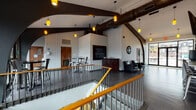 This dynamic fitness facility is defined by its striking architectural design, featuring soaring vaulted ceilings with bold dark curved beam accents and white shiplap wall paneling throughout. Warm amber and soft pendant lighting adds an inviting glow across the space.

The centerpiece is a generously sized open studio with gleaming hardwood floors, floor-to-ceiling glass windows along one wall that flood the room with natural light and offer views of the outdoors. The studio is equipped with a hanging heavy bag and a speed bag mount, making it well-suited for a variety of fitness classes and training sessions.

The lounge and reception area features large-format tile flooring, a dark wood credenza with a framed chalkboard display for scheduling, and a decorative sunburst mirror on the shiplap wall. Plush dark leather armchairs provide a comfortable space for members to relax.

A dedicated refreshment bar area includes a row of industrial-style metal barstools along a counter, with a wall-mounted television above and small appliances nearby. Additional gym equipment is visible in an adjacent workout zone. A staircase with a warm wood handrail and metal balusters connects different areas of the facility, completing this well-appointed, modern fitness environment.