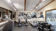 Located at 300 Springboro Lane in Columbus, Ohio, this fitness center offers a well-equipped, single-floor exercise facility designed to support a comprehensive workout experience. The space features vaulted ceilings with distinctive exposed wood beams, adding architectural character to the room, while track lighting illuminates the area throughout. A textured ceiling and neutral wall tones create a clean, functional atmosphere. Large windows along the walls allow natural light to filter in, contributing to an open and energizing environment. The facility is outfitted with a variety of cardio equipment, including treadmills and elliptical machines, positioned to take advantage of the room's layout. A dedicated dumbbell rack provides a range of free weight options for strength training. Full-length mirrors line one wall, enhancing the sense of space and allowing users to monitor their form during workouts. Dark rubber flooring covers the ground, offering durability and safety underfoot. A mounted television and posted fitness rules round out the practical amenities. The overall ambiance is practical and motivating, making this fitness center a well-rounded resource for residents seeking convenient access to exercise facilities.