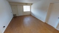 This single-floor space presents a clean, open layout designed for functional everyday living. The main living area is highlighted by warm-toned hardwood-style flooring that extends throughout, lending a cohesive and inviting feel to the room. Smooth, neutral-painted walls in a soft beige tone create a bright, understated backdrop that complements a variety of interior styles.

A front entry door with a classic six-panel design serves as the main point of entry, positioned alongside a window that allows natural light to filter into the space. The window provides a modest connection to the outdoors while keeping the interior well-lit during daylight hours. Practical details include floor-level heating and cooling vents, ensuring year-round climate control throughout the unit.

The open floor plan allows for flexible furniture arrangement, making it easy to define living and other functional zones according to individual needs. White baseboards trim the perimeter of the room, adding a subtle finishing detail that contrasts neatly against the flooring. Overall, the space offers a straightforward, move-in-ready environment with a neutral palette suited to a range of personal styles.