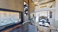 This single-story community clubhouse makes a striking first impression with its dramatic double-height ceilings that soar above the main lounge and entry areas. Large clerestory windows draw in abundant natural light, illuminating the polished large-format tile flooring that extends throughout the space. The main lounge area is anchored by a sculptural, floor-to-ceiling geometric bookshelf wall with integrated lighting and a sleek linear fireplace set against a marble-inspired surround, creating an inviting yet architecturally bold focal point. Plush seating arrangements, including leather sofas, upholstered ottomans, and accent chairs, are thoughtfully arranged on striped area rugs to define conversation zones.

An adjacent lounge area features deeply textured wall treatments, built-in niche seating, and a tall framed art installation, adding an intimate, gallery-like quality. A full kitchen and bar area is outfitted with dark, flat-front cabinetry with brass hardware, a herringbone tile backsplash, stainless steel appliances, and a generous island with upholstered bar seating beneath a row of striking brass cylindrical pendant lights. Wide corridors with polished concrete-style flooring and curated wall art connect each area, while a sliding barn-style door adds a refined industrial detail throughout this cohesive, upscale communal setting.
