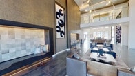 This single-story community clubhouse makes a striking first impression with its dramatic double-height ceilings that soar above the main lounge and entry areas. Large clerestory windows draw in abundant natural light, illuminating the polished large-format tile flooring that extends throughout the space. The main lounge area is anchored by a sculptural, floor-to-ceiling geometric bookshelf wall with integrated lighting and a sleek linear fireplace set against a marble-inspired surround, creating an inviting yet architecturally bold focal point. Plush seating arrangements, including leather sofas, upholstered ottomans, and accent chairs, are thoughtfully arranged on striped area rugs to define conversation zones.

An adjacent lounge area features deeply textured wall treatments, built-in niche seating, and a tall framed art installation, adding an intimate, gallery-like quality. A full kitchen and bar area is outfitted with dark, flat-front cabinetry with brass hardware, a herringbone tile backsplash, stainless steel appliances, and a generous island with upholstered bar seating beneath a row of striking brass cylindrical pendant lights. Wide corridors with polished concrete-style flooring and curated wall art connect each area, while a sliding barn-style door adds a refined industrial detail throughout this cohesive, upscale communal setting.