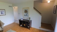 This three-story home offers a thoughtfully designed layout spread across multiple levels, connected by a staircase with a sleek dark handrail. The entryway sets a welcoming tone, featuring warm hardwood floors that extend throughout the main level, creating a cohesive and inviting atmosphere. Soft, neutral wall tones in a muted sage-green hue lend the space a calm, understated character that complements the natural wood elements. A console unit with open shelving provides practical storage near the entry, adding both function and subtle decorative interest. Adjacent to the staircase, a hallway leads further into the home, hinting at additional rooms beyond. Natural light filters into the space, enhancing the airy feel of the interior. The staircase, framed by a clean white enclosure, rises to the upper floors, suggesting well-defined living zones distributed across the home's three levels. Overall, the space presents a clean, classic aesthetic with a balance of practicality and comfort throughout.