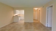This single-floor space is designed with an open, functional layout that promotes a sense of flow between living areas. The main living area features warm-toned ceramic tile flooring that extends throughout, providing a cohesive and easy-to-maintain surface underfoot. Neutral beige walls create a clean, versatile backdrop suitable for a variety of décor styles.

The kitchen is accessible directly from the living area via an open archway, making it a natural extension of the main space. It is outfitted with white cabinetry offering ample storage, white appliances including a range and dishwasher, and a peninsula counter that serves as a natural divider between the kitchen and living area. The light-colored countertops complement the overall bright, airy aesthetic of the kitchen.

A hallway visible from the living area leads to additional rooms, with a closet featuring a double-door configuration adding practical storage. The space is finished with clean lines and a consistent color palette throughout, contributing to a bright and welcoming atmosphere. The straightforward, single-level layout makes the space highly accessible and easy to navigate.