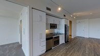 This contemporary single-floor space showcases a clean, modern aesthetic designed for both functionality and style. The kitchen serves as a focal point, outfitted with a full suite of stainless steel appliances including a refrigerator, range with double oven, built-in microwave, and a gas cooktop with a dark countertop surface. An abundance of floor-to-ceiling white cabinetry with sleek bar-pull hardware provides generous storage, keeping the space organized and visually uncluttered. Track lighting runs along the ceiling, offering adjustable, directed illumination throughout the kitchen and adjoining areas. The flooring features rich, dark hardwood-style planks that extend seamlessly through the space, adding warmth and contrast against the bright white cabinetry and light-toned walls. The layout is thoughtfully open, with a hallway visible in the background leading to additional areas of the unit. Ventilation is well-integrated with ceiling vents maintaining a tidy, modern finish. Overall, the space exudes a minimalist, urban character with a practical and polished design sensibility suited for modern living.