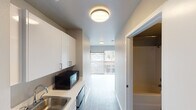 This single-floor unit offers a clean, modern design with a thoughtfully efficient layout that makes great use of every square foot. The space is finished with light wood-tone flooring throughout the main living area, complemented by crisp white walls and flush ceiling light fixtures that maintain a bright, contemporary aesthetic.

The kitchenette runs along one wall and is well-appointed with white upper and lower cabinetry, a stainless steel sink, a built-in microwave, and additional appliances, all set against a clean white countertop. The galley-style arrangement keeps everything within easy reach while maintaining an open sightline toward the living area.

The main living space is open and airy, benefiting from abundant natural light streaming through a large sliding glass door. This door leads to a private balcony with views of a lively urban streetscape, offering a pleasant connection to the surrounding neighborhood. A spacious closet provides ample built-in storage, keeping the living area uncluttered.

The entry hallway features dark hardwood-style flooring that transitions neatly into the broader living space, adding a subtle contrast to the overall palette. The unit's neutral tones, modern finishes, and practical design make it a versatile and comfortable space well-suited for everyday living.