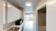 This single-floor unit offers a clean, modern design with a thoughtfully efficient layout that makes great use of every square foot. The space is finished with light wood-tone flooring throughout the main living area, complemented by crisp white walls and flush ceiling light fixtures that maintain a bright, contemporary aesthetic.

The kitchenette runs along one wall and is well-appointed with white upper and lower cabinetry, a stainless steel sink, a built-in microwave, and additional appliances, all set against a clean white countertop. The galley-style arrangement keeps everything within easy reach while maintaining an open sightline toward the living area.

The main living space is open and airy, benefiting from abundant natural light streaming through a large sliding glass door. This door leads to a private balcony with views of a lively urban streetscape, offering a pleasant connection to the surrounding neighborhood. A spacious closet provides ample built-in storage, keeping the living area uncluttered.

The entry hallway features dark hardwood-style flooring that transitions neatly into the broader living space, adding a subtle contrast to the overall palette. The unit's neutral tones, modern finishes, and practical design make it a versatile and comfortable space well-suited for everyday living.