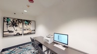 Located at 4735 Sepulveda Blvd in Los Angeles, California, this single-floor business center is a well-appointed, efficiently designed workspace suited for professional productivity. The room features clean white walls and recessed lighting that creates a bright, focused atmosphere. A sleek, dark gray desk runs along the wall and is outfitted with an HP all-in-one desktop computer, a multifunction printer, a full keyboard, and mouse — providing all essential tools for printing, scanning, and computing needs. Two black leather ergonomic chairs offer comfortable seating for multiple users. The floor is covered in a bold, contemporary patterned carpet in shades of teal, black, and white, adding a vibrant design element that reflects the creative energy of the Los Angeles locale. A standout decorative feature is a large diptych wall art piece depicting a vintage VW van among palm trees, evoking a classic Southern California coastal aesthetic. A tall dark storage cabinet provides additional organizational space. The room is accessed through a glass-paneled door that allows visibility into the adjacent corridor. Safety features include ceiling-mounted smoke detectors and a fire sprinkler system, ensuring a secure working environment.