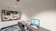 Located at 4735 Sepulveda Blvd in Los Angeles, California, this single-floor business center is a well-appointed, efficiently designed workspace suited for professional productivity. The room features clean white walls and recessed lighting that creates a bright, focused atmosphere. A sleek, dark gray desk runs along the wall and is outfitted with an HP all-in-one desktop computer, a multifunction printer, a full keyboard, and mouse — providing all essential tools for printing, scanning, and computing needs. Two black leather ergonomic chairs offer comfortable seating for multiple users. The floor is covered in a bold, contemporary patterned carpet in shades of teal, black, and white, adding a vibrant design element that reflects the creative energy of the Los Angeles locale. A standout decorative feature is a large diptych wall art piece depicting a vintage VW van among palm trees, evoking a classic Southern California coastal aesthetic. A tall dark storage cabinet provides additional organizational space. The room is accessed through a glass-paneled door that allows visibility into the adjacent corridor. Safety features include ceiling-mounted smoke detectors and a fire sprinkler system, ensuring a secure working environment.
