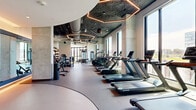 Located at 30 Peabody St in Nashville, Tennessee, this state-of-the-art fitness center spans a generously sized single floor, offering a comprehensive and well-appointed wellness facility. The space is defined by its striking architectural design, featuring a combination of exposed concrete ceilings, warm wood slat accents, and dramatic hexagonal LED lighting fixtures that create a modern, energetic ambiance throughout.

Large floor-to-ceiling windows line much of the facility, flooding the interior with abundant natural light and providing sweeping views of the surrounding Nashville cityscape. The main workout floor is divided into distinct training zones: a turf area equipped with a functional training rig, TRX suspension systems, plyo boxes, and rowing machines; a dedicated cardio zone lined with premium TRUE treadmills, stair climbers, and ellipticals; and a strength training area housing selectorized machines, free weights, and a Smith machine with bench press stations.

A relaxation room features massage chairs set against textured stone-look wall panels with expansive city-facing windows. The facility also includes a sauna, a refreshment station with built-in storage and a beverage refrigerator, and glass-partitioned entryways that enhance the open, airy feel. Rubber flooring and mirrored walls complete the polished, professional atmosphere of this exceptional fitness destination.